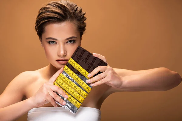 Young woman with chocolate bar in yellow measuring tape isolated on beige — Stock Photo