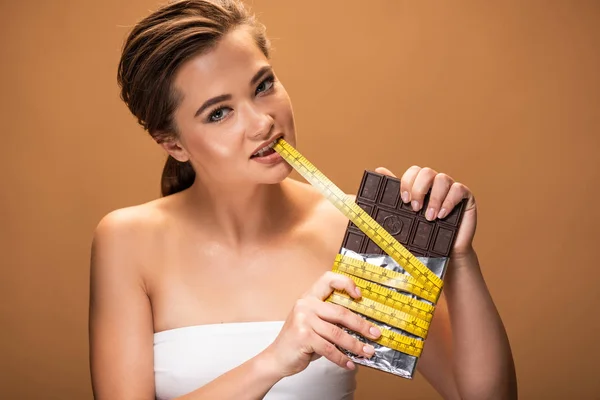 Young woman with yellow measuring tape in mouth holding chocolate bar isolated on beige — Stock Photo