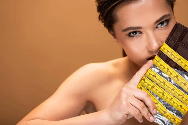 Giovane donna nuda con barretta di cioccolato in nastro di misurazione giallo isolato su beige — Foto stock