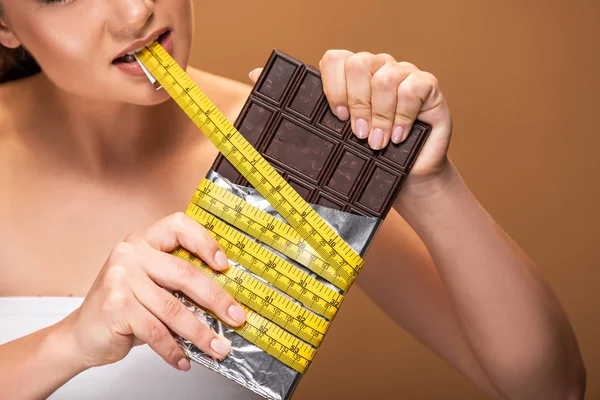 Partial view of young woman holding yellow measuring tape in mouth and chocolate bar isolated on beige — Stock Photo