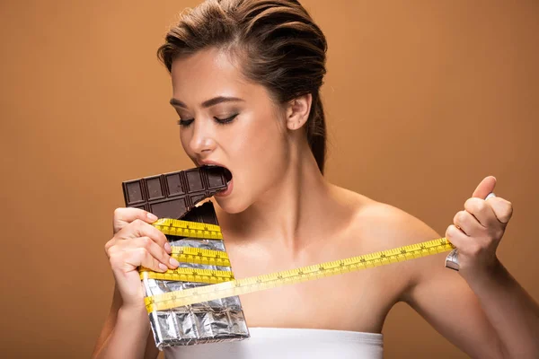 Young woman eating chocolate bar in yellow measuring tape isolated on beige — Stock Photo