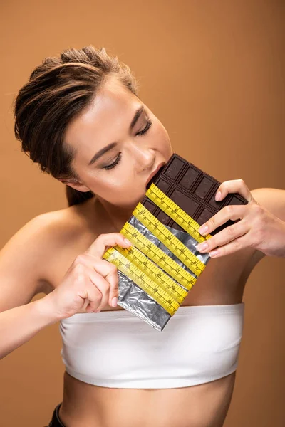 Young woman with yellow measuring tape eating chocolate isolated on beige — Stock Photo