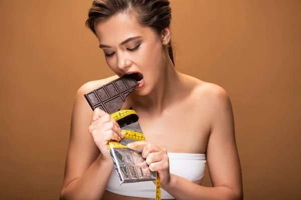 Joven mujer delgada con cinta métrica amarilla mordiendo barra de chocolate aislado en beige - foto de stock