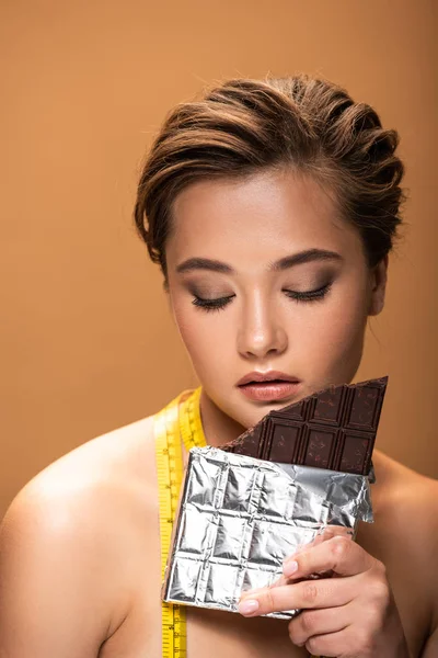 Naked young woman with closed eyes and yellow measuring tape holding chocolate bar isolated on beige — Stock Photo