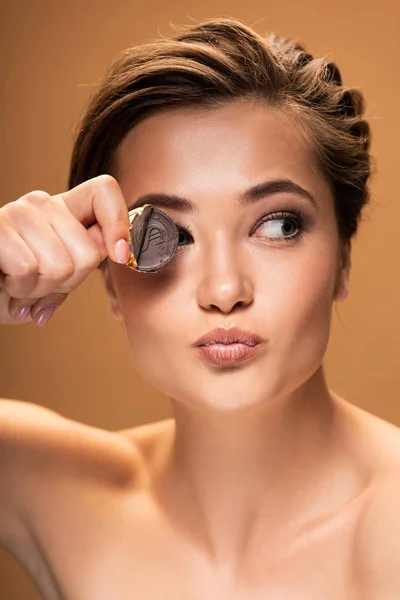 Naked young woman holding chocolate coin in golden foil near eye and looking away isolated on beige — Stock Photo