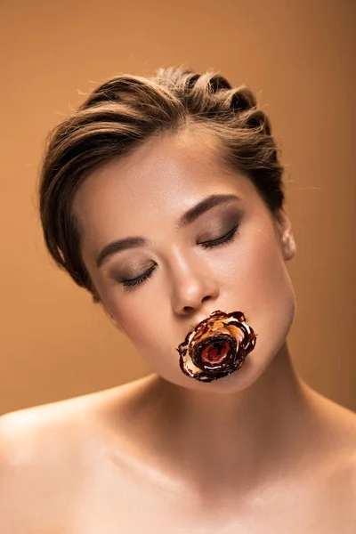 Обнаженная женщина с закрытыми глазами, держащая розу во рту, покрытую расплавленным шоколадом, изолированным на бежевом — стоковое фото