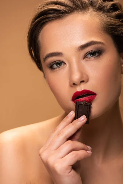 Attractive young woman holding chocolate piece near mouth isolated on beige — Stock Photo