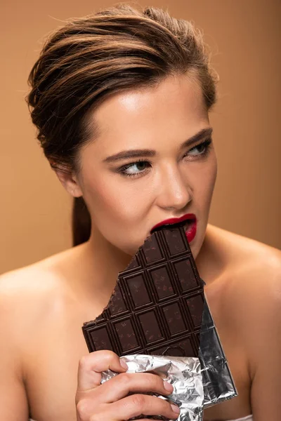 Красива гола жінка з червоними губами кусає шоколадний батончик у срібній фользі ізольована на бежевому — стокове фото