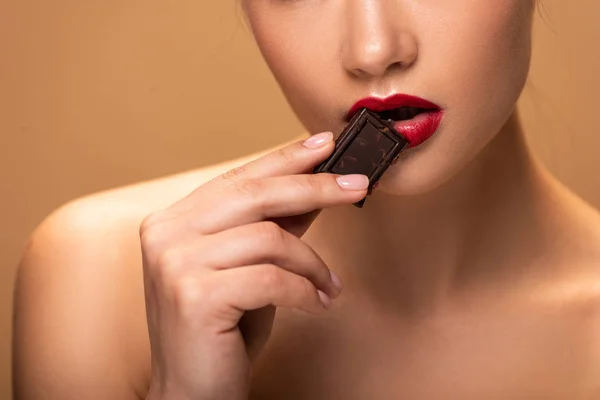 Обрезанный вид обнаженной женщины, держащей шоколадный кусочек возле губ, изолированных на бежевый — стоковое фото