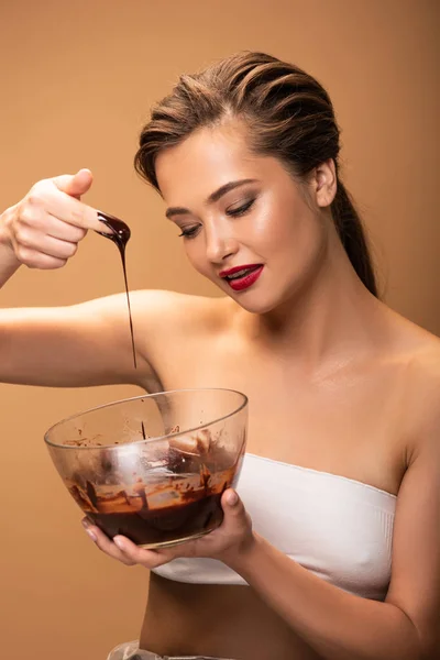 Beautiful woman with red lips holding melted chocolate on finger above bowl isolated on beige — Stock Photo