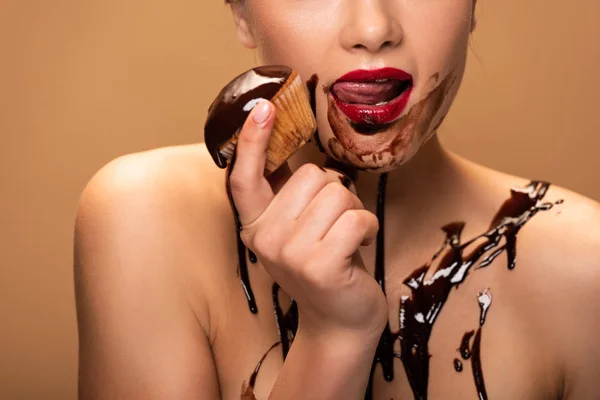 Partial view of naked woman licking red lips with chocolate spills on skin and holding muffin isolated on beige — Stock Photo