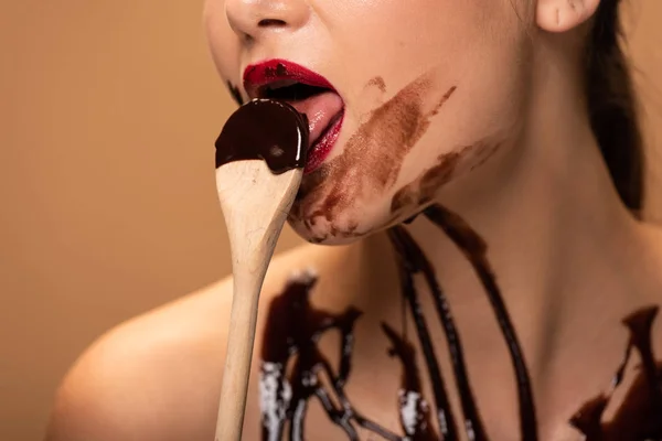 Vista recortada de mujer desnuda con labios rojos y derrames de chocolate en cuchara lamiendo la piel aislada en beige - foto de stock