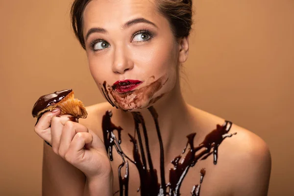 Hermosa mujer desnuda de ensueño con labios rojos y derrames de chocolate en la piel comiendo muffin aislado en beige — Stock Photo