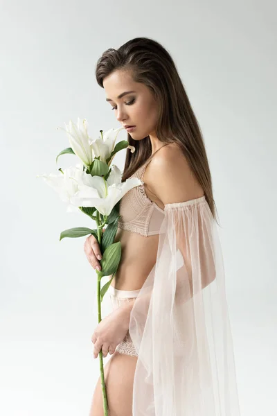 Zarte junge Frau in beiger Dessous und Netzärmeln, die Lilien isoliert auf weißem Grund betrachtet — Stockfoto