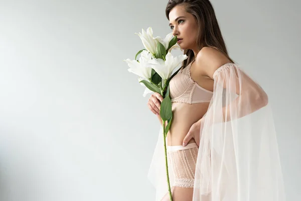 Side view of delicate young woman in beige lingerie and mesh sleeves smelling lilies isolated on white — Stock Photo