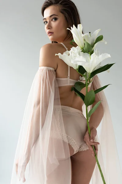 Low angle view of delicate young woman in beige lingerie and mesh sleeves holding bouquet of lilies behind back isolated on white — Stock Photo