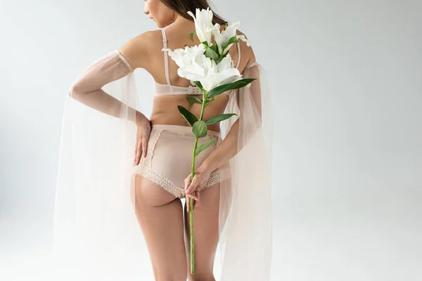 Vue arrière de la jeune femme délicate en lingerie beige et manches en mesh tenant bouquet de lis isolés sur blanc — Photo de stock