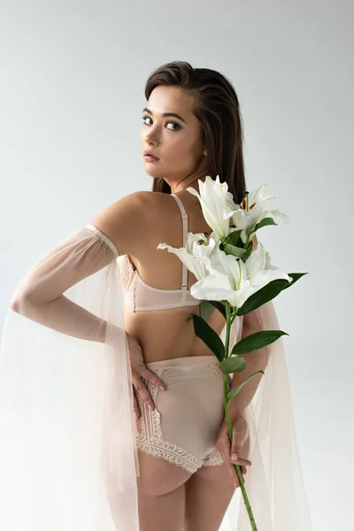 Tendre jeune femme en lingerie beige et manches en mesh tenant des lys derrière le dos isolé sur blanc — Photo de stock