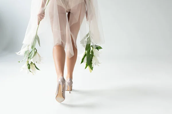 Rückansicht einer jungen Frau in beiger Dessous und Netzärmeln, die mit Lilien auf weißem Hintergrund spazieren geht — Stockfoto