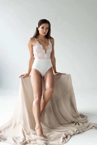 Tender young woman in white lacy bodysuit sitting on beige cloth isolated on white — Stock Photo
