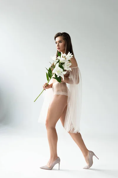 Side view of tender young woman in beige lingerie and mesh sleeves walking with white lilies — Stock Photo