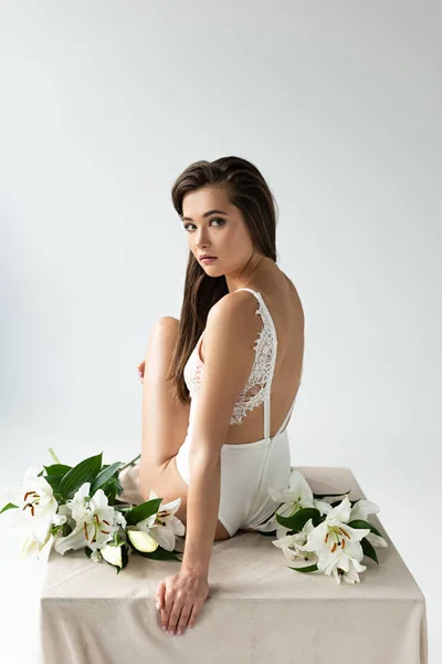 Zarte junge Frau im weißen Spitzenbody sitzt inmitten von Lilien isoliert auf weißem Grund — Stockfoto