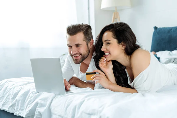 Усміхнена молода пара використовує ноутбук і тримає кредитну картку в ліжку, інтернет-покупки — стокове фото