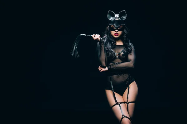 Sexy giovane bruna donna in costume bdsm e maschera con frusta fustigazione in pelle isolato su nero — Foto stock