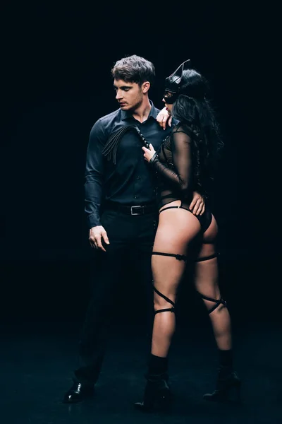 Joven hombre y sexy morena mujer en bdsm traje con azotes látigo en negro - foto de stock