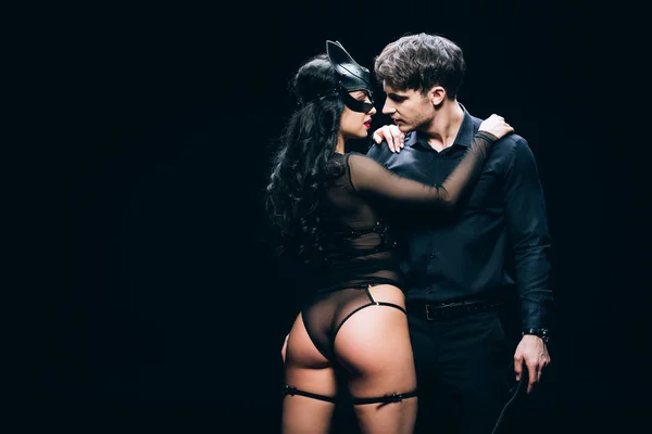 Junger Mann steht und schaut sexy brünette Frau in Maske und BDSM-Kostüm isoliert auf schwarz an — Stockfoto