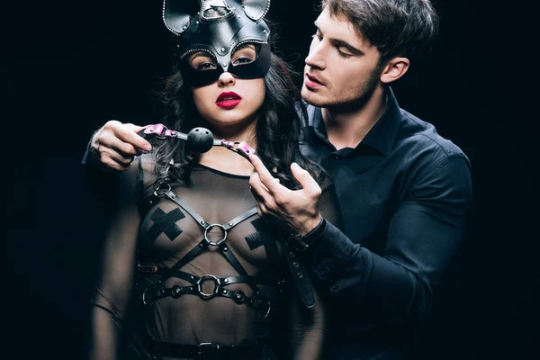 Joven hombre sosteniendo mordaza cerca sexy mujer en máscara y bdsm traje aislado en negro - foto de stock