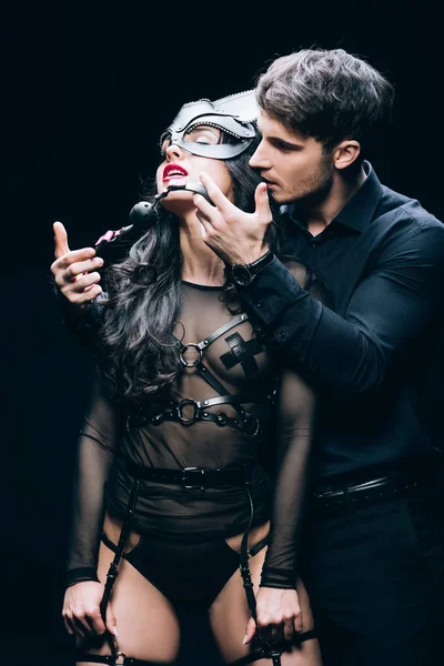 Handsome young man holding gag near sexy passionate woman in mask and bdsm costume isolated on black — Stock Photo
