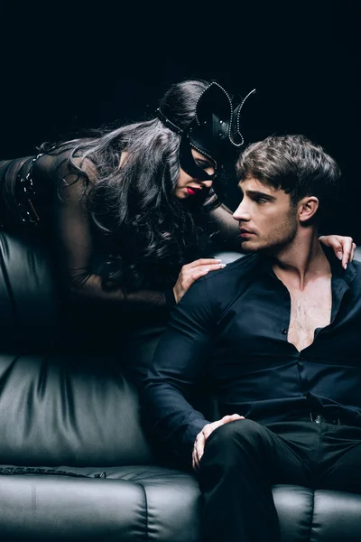 Donna sexy in maschera bdsm spogliarsi fidanzato sul divano in pelle nera isolato su nero — Foto stock