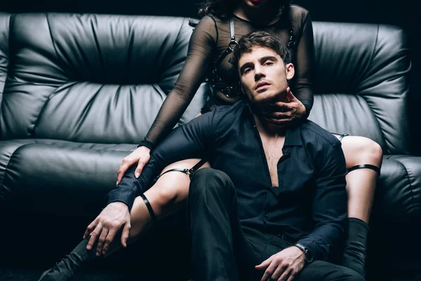 Leidenschaftlicher schöner Mann sitzt neben sexy Frau im BDSM-Kostüm auf schwarzem Ledersofa — Stockfoto