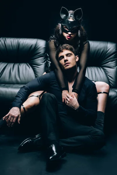 Sexy donna in costume bdsm e maschera abbracciando l'uomo sul divano in pelle nera isolato su nero — Foto stock