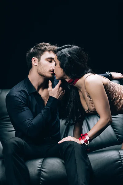 Sexy morena mulher no bege roupa interior e vermelho algemas beijando homem no couro sofá isolado no preto — Fotografia de Stock