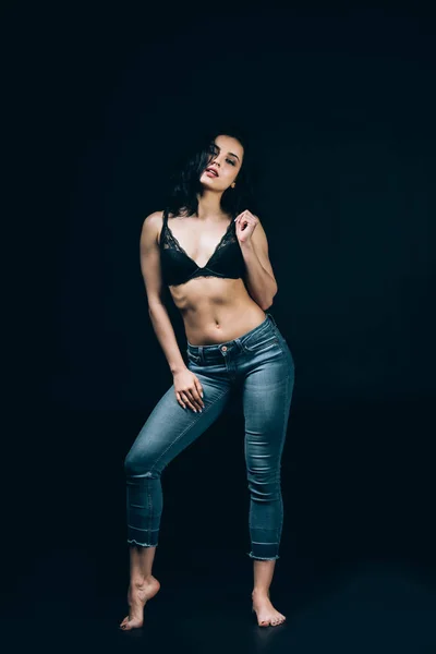 Sexy brunette barefoot woman in blue jeans and bra posing isolated on black — Stock Photo