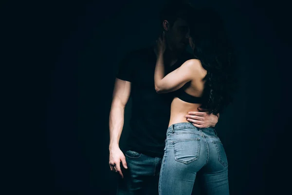 Мужчина целует девушку в бюстгальтере и джинсах, изолированных на черном — стоковое фото