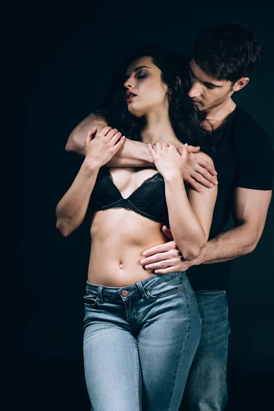 Handsome young man embracing undressed girl in bra isolated on black — Stock Photo