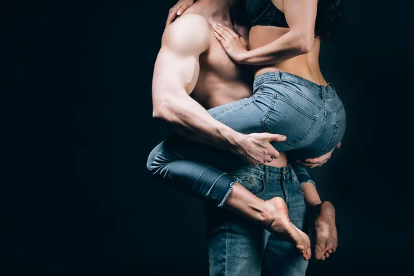 Corte vista de homem em jeans com muscular torso segurando namorada isolado no preto — Fotografia de Stock