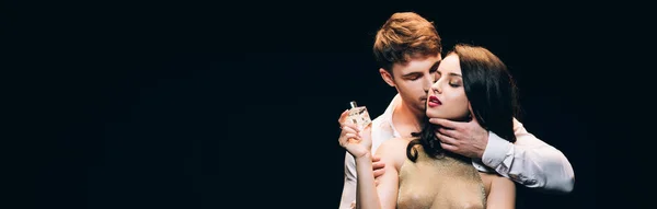 Panoramic shot of young brunette woman holding perfume while boyfriend kissing her isolated on black — Stock Photo