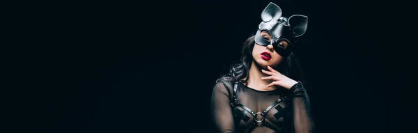 Panoramic shot of sexy brunette woman in bdsm costume and mask isolated on black — Stock Photo