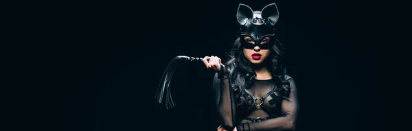 Panoramic shot of sexy young brunette woman in bdsm costume and mask with leather flogging whip isolated on black — Stock Photo