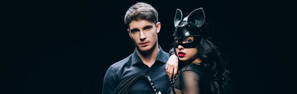 Plano panorámico de hombre joven y mujer morena sexy en traje de bdsm con látigo azotes aislado en negro - foto de stock