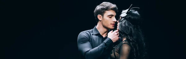 Plano panorámico de hombre joven besar sexy morena mujer en bdsm traje aislado en negro - foto de stock