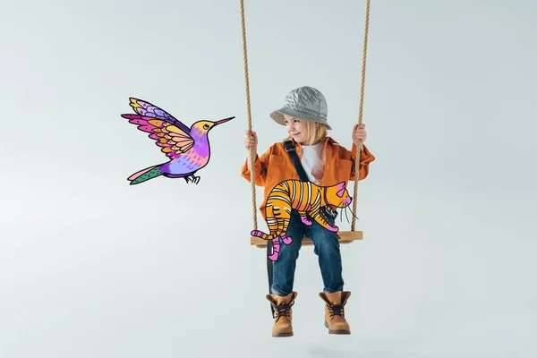 Mignon enfant en jeans et chemise orange assis sur balançoire avec chat fantaisie sur les genoux et en regardant l'illustration d'oiseau coloré sur fond gris — Photo de stock