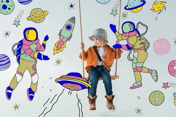 Carino bambino in jeans e camicia arancione seduto su altalena e guardando spazio fata con astronauti illustrazione su sfondo grigio — Foto stock