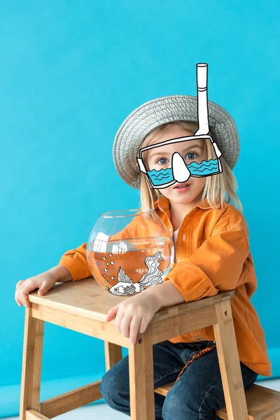 Kid in silver hat, orange shirt and snorkel sitting on stairs with fairy fish in aquarium on blue background — Stock Photo