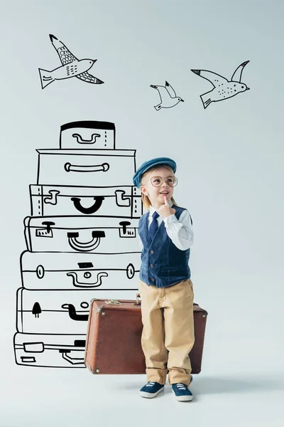 Enfant rêveur en gilet rétro et casquette tenant la valise et regardant loin sur fond gris avec des sacs de voyage et des oiseaux volants illustration fantaisie — Photo de stock