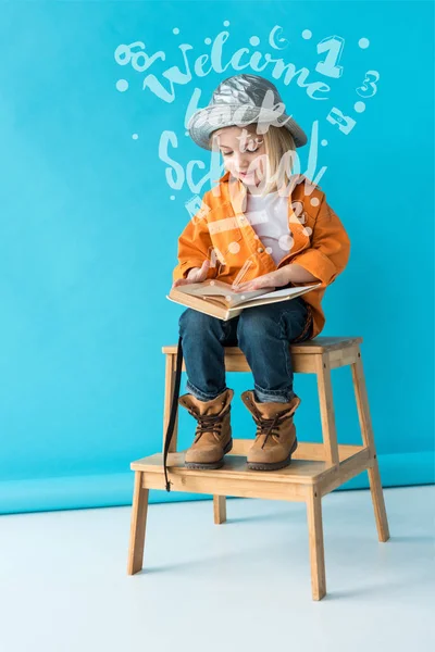 Kid in jeans and orange shirt sitting on stairs and reading book on blue background with welcome back to school lettering — Stock Photo